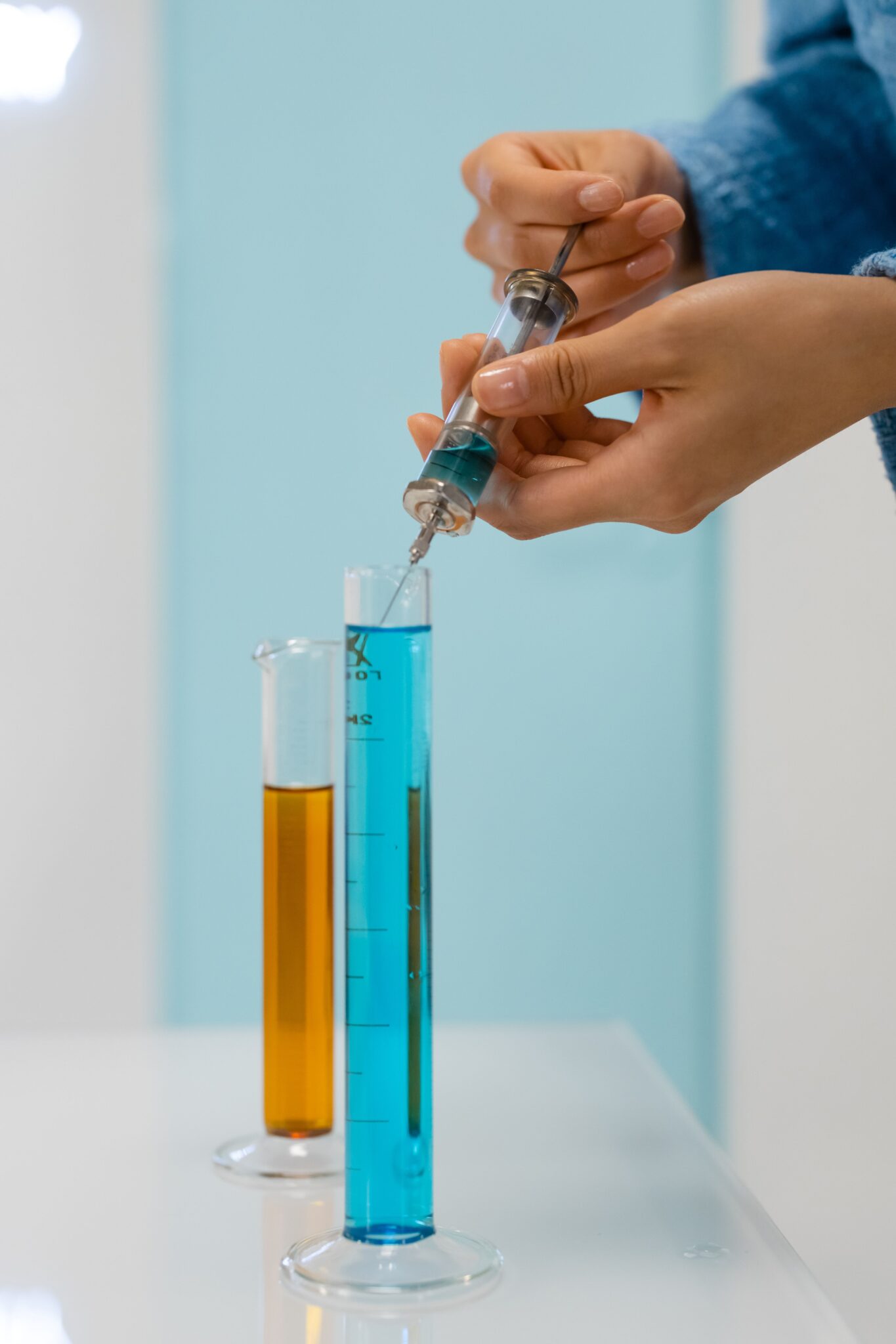 Close-up of hands using a syringe in a lab with colorful liquids in graduated cylinders.